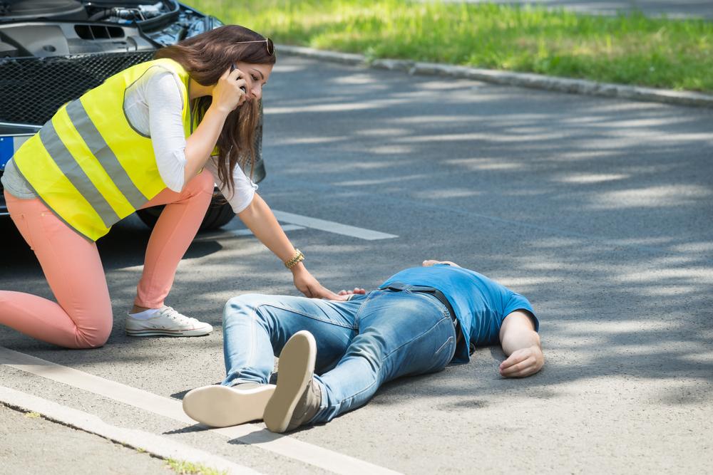 crossing guard calling emergency services with man in blue shirt laying on the ground after being struck by a car.