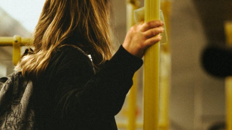 A woman with a backpack holds a yellow grab pole while riding public transit